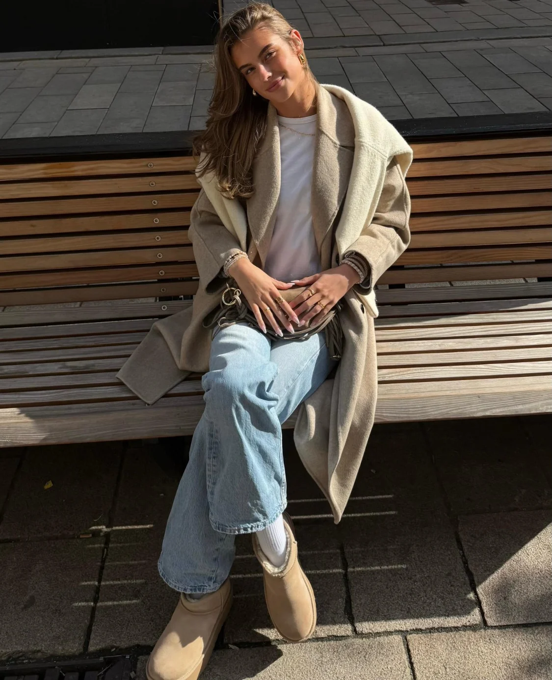 Jule im beigen Mantel und Jeans auf einer Holzbank in der Sonne